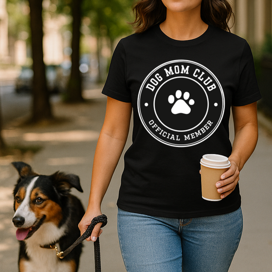 Woman wearing a 'Dog Mom Club' t-shirt holding a coffee cup and walking a dog on a sidewalk.